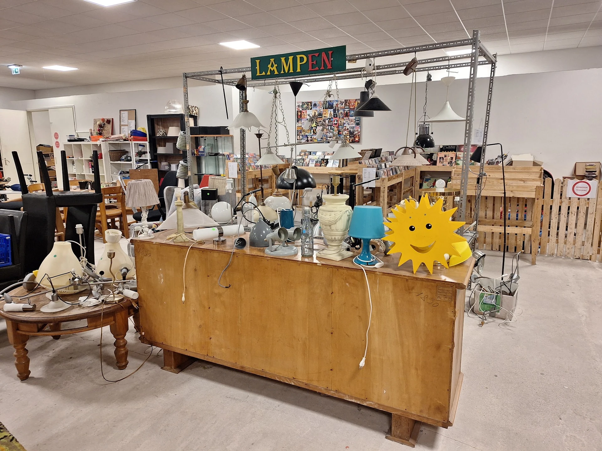 Houten kast met huishoudelijke spullen, lampen en decoratie; achter een metalen frame met 'LAMPEN' tekstbord