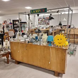 Houten kast met huishoudelijke spullen, lampen en decoratie; achter een metalen frame met 'LAMPEN' tekstbord