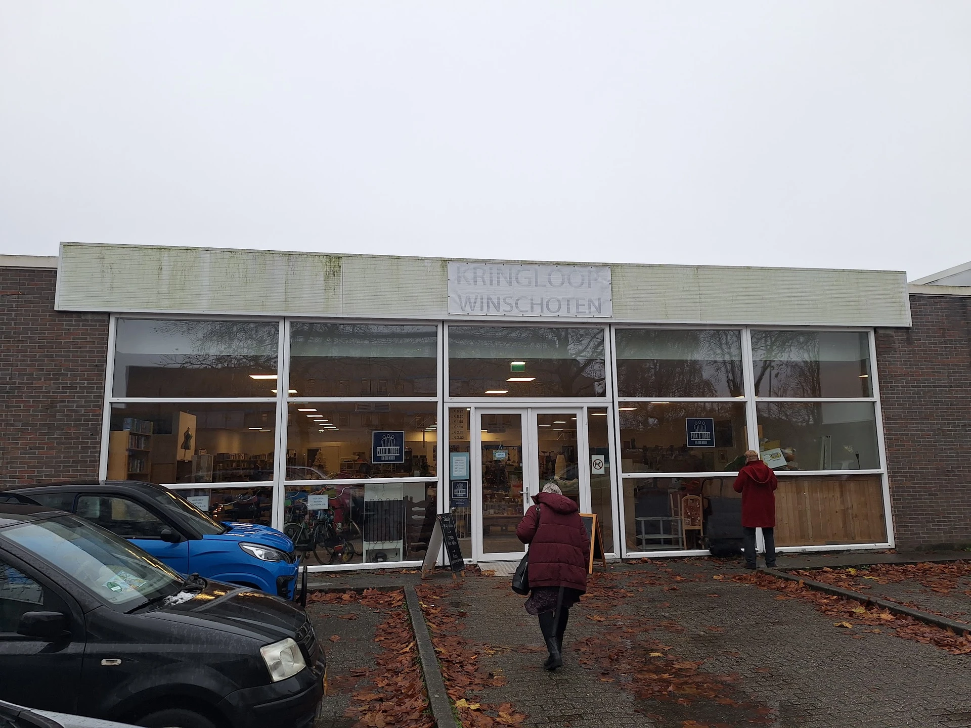 Voorgevel van kringloopwinkel met grote ramen, enkele bezoekers voor de ingang, geparkeerde auto's en herfstbladeren op het p