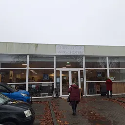 Voorgevel van kringloopwinkel met grote ramen, enkele bezoekers voor de ingang, geparkeerde auto's en herfstbladeren op het p