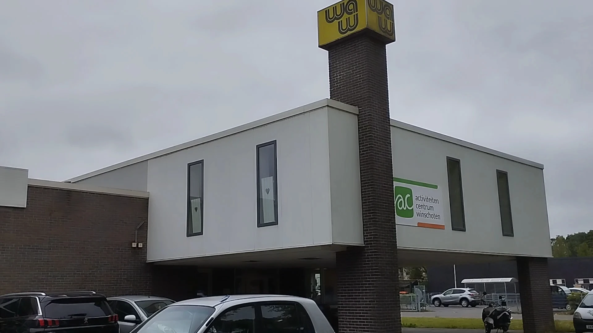 Moderne gevel van kringloopwinkel met witte bekleding, bakstenen schouw, gele logo en groene huismerksticker