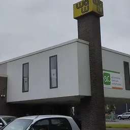 Moderne gevel van kringloopwinkel met witte bekleding, bakstenen schouw, gele logo en groene huismerksticker