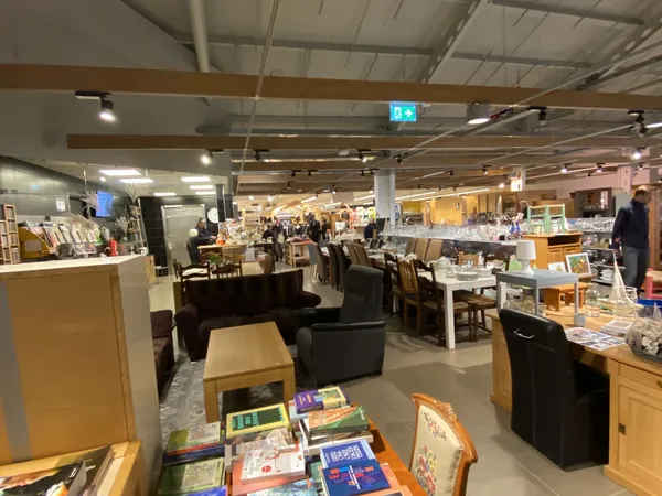 Ruim kringloopwinkelinterieur met boeken op tafels, zitmeubels, stellingen met spullen en bezoekers in de winkel