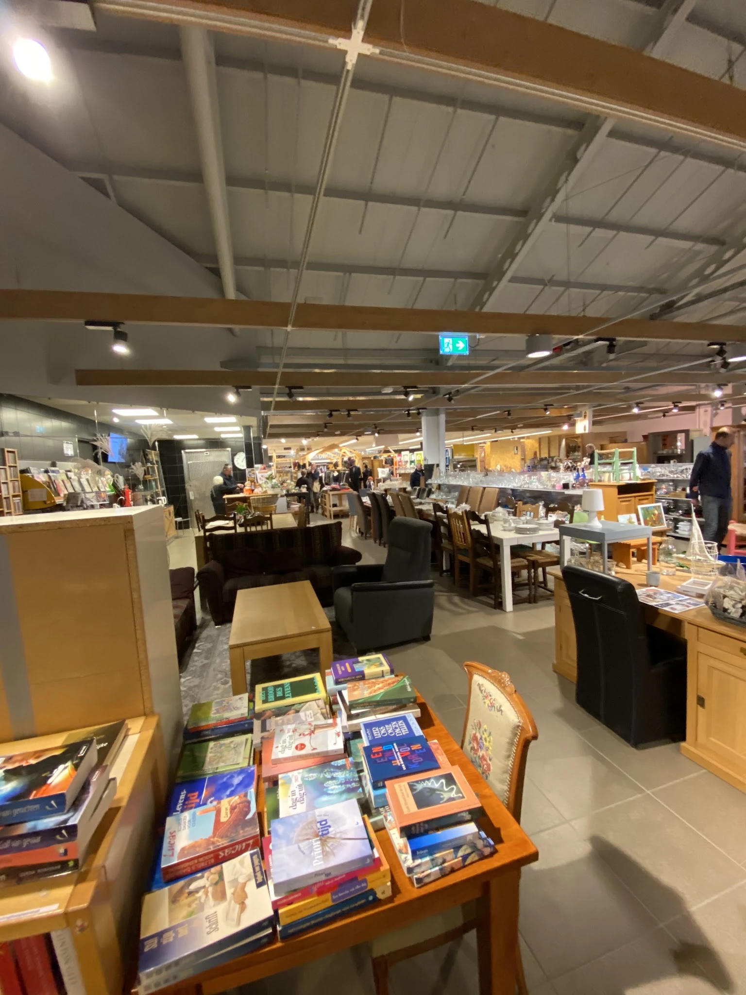 Ruim kringloopwinkelinterieur met boeken op tafels, zitmeubels, stellingen met spullen en bezoekers in de winkel