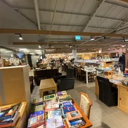 Ruim kringloopwinkelinterieur met boeken op tafels, zitmeubels, stellingen met spullen en bezoekers in de winkel