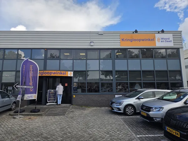 Gevel van kringloopwinkel met oranje logo, grote ramen, parkeerplaats met auto's ervoor en blauwe banner aan zijkant