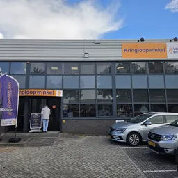 Gevel van kringloopwinkel met oranje logo, grote ramen, parkeerplaats met auto's ervoor en blauwe banner aan zijkant