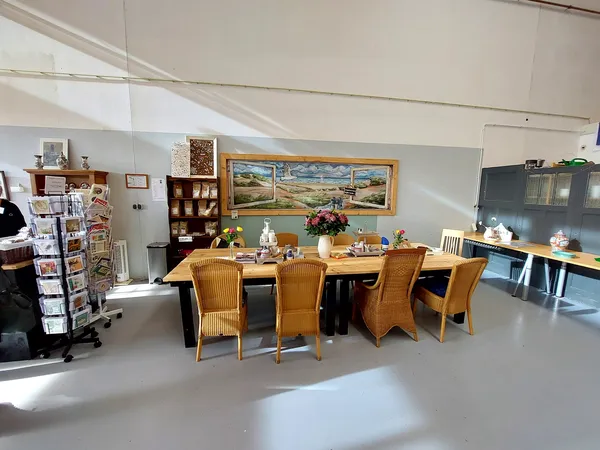 Winkelvloer met eettafel, stoelen en schilderij aan de muur, stellingen met spullen op de achtergrond
