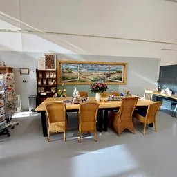 Winkelvloer met eettafel, stoelen en schilderij aan de muur, stellingen met spullen op de achtergrond