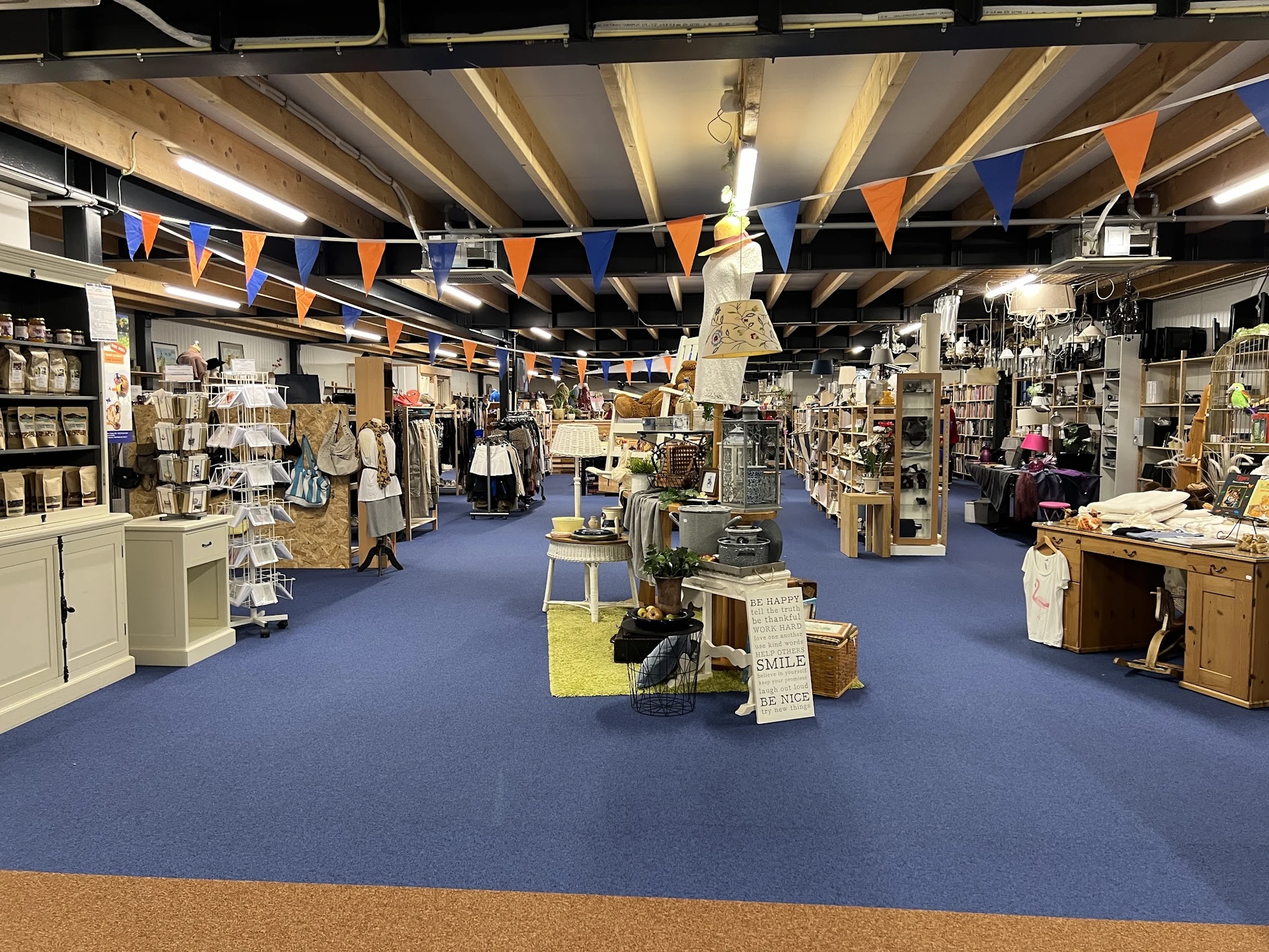Overzicht van de winkelvloer met blauwe vloer, stellingen, displays en diverse tweedehands artikelen verspreid door de winkel
