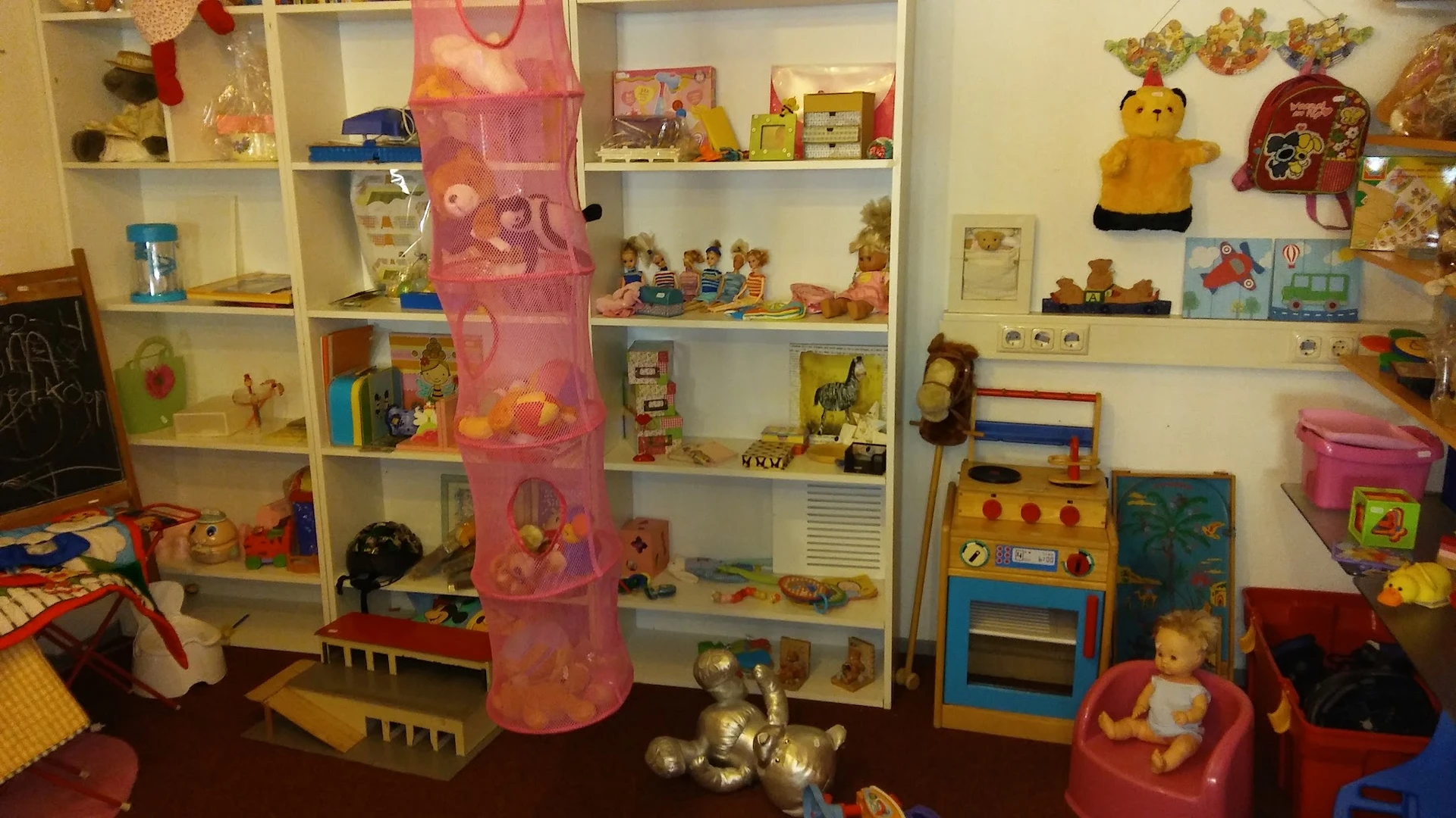 Speelgoed en kinderspeelgoed uitgestald op witte stellingen en op de vloer, waaronder poppen, knuffels, speelkeuken en een ro