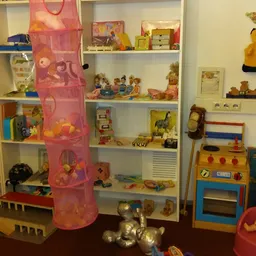 Speelgoed en kinderspeelgoed uitgestald op witte stellingen en op de vloer, waaronder poppen, knuffels, speelkeuken en een ro