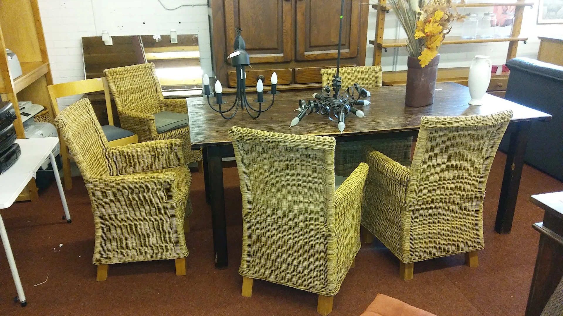 Eettafel met zes rieten stoelen met armleuningen en houten poten, versierd met kandelaar en bloemen.