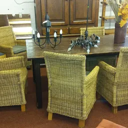 Eettafel met zes rieten stoelen met armleuningen en houten poten, versierd met kandelaar en bloemen.
