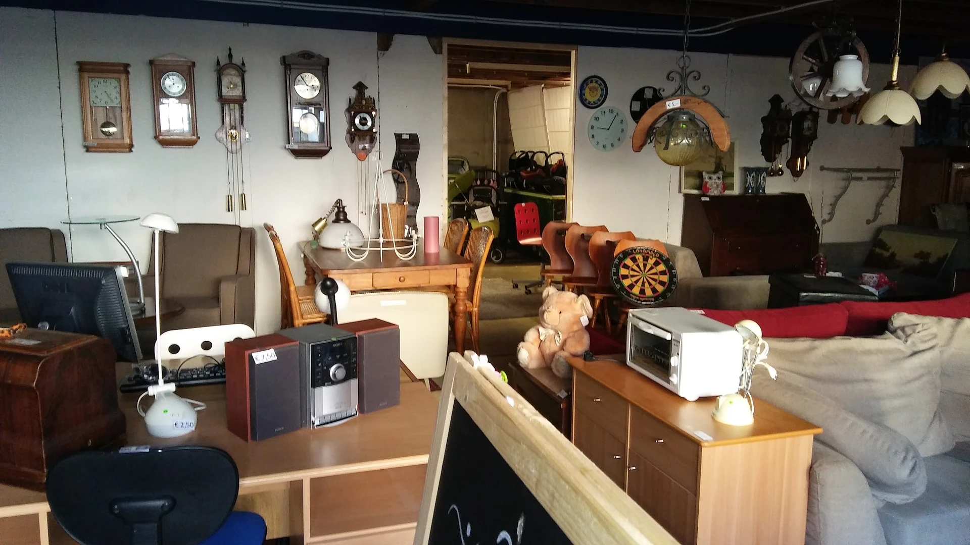 Kringloopwinkelinterieur met tafels, stoelen, klokken aan de wand, dartbord en diverse huishoudelijke artikelen