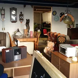 Kringloopwinkelinterieur met tafels, stoelen, klokken aan de wand, dartbord en diverse huishoudelijke artikelen