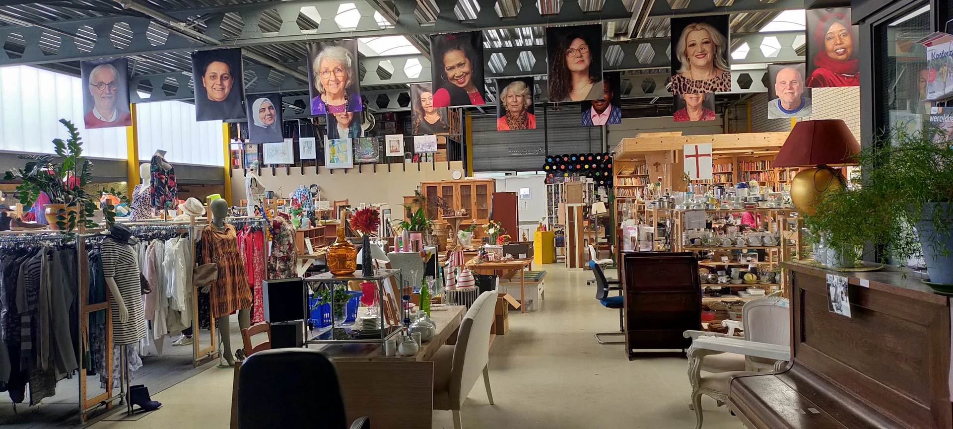 Ruim interieur van kringloopwinkel met kledingrekken, tafels met huishoudartikelen, boeken en diverse spullen; portretten van