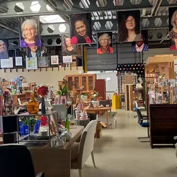 Ruim interieur van kringloopwinkel met kledingrekken, tafels met huishoudartikelen, boeken en diverse spullen; portretten van