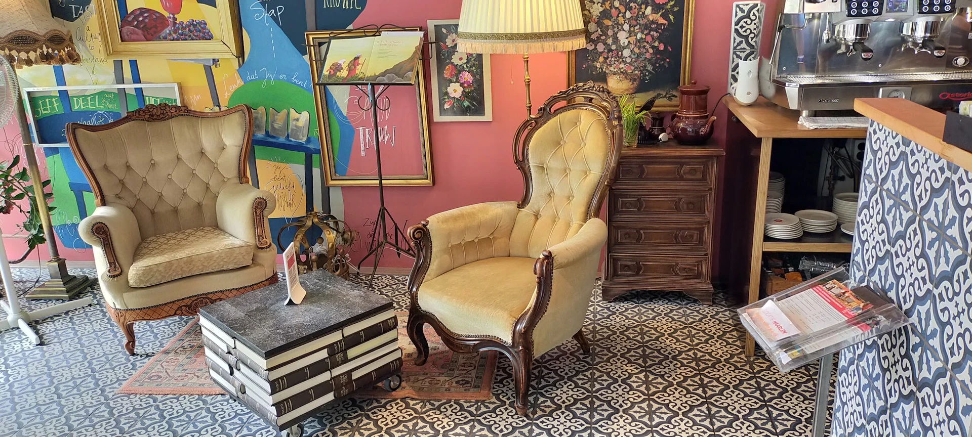 Vintage fauteuil met gele bekleding en houten armleuningen, omgeven door boeken, kunstwerk en antieke meubels in een kringloo