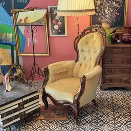 Vintage fauteuil met gele bekleding en houten armleuningen, omgeven door boeken, kunstwerk en antieke meubels in een kringloo