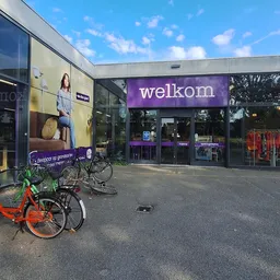 Gevel van kringloopwinkel met paarse welkom-bord, grote glazen puien en twee fietsen geparkeerd ervoor