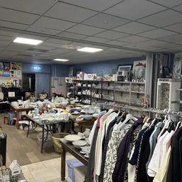 Winkelvloer met kledingrek, tafels met diverse spullen en stellingen tegen de muur in kringloopwinkel