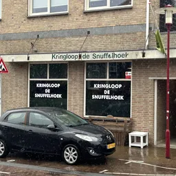 Voorgevel van kringloopwinkel met zwarte etalageruiten en winkelnaam op bord, auto geparkeerd ervoor