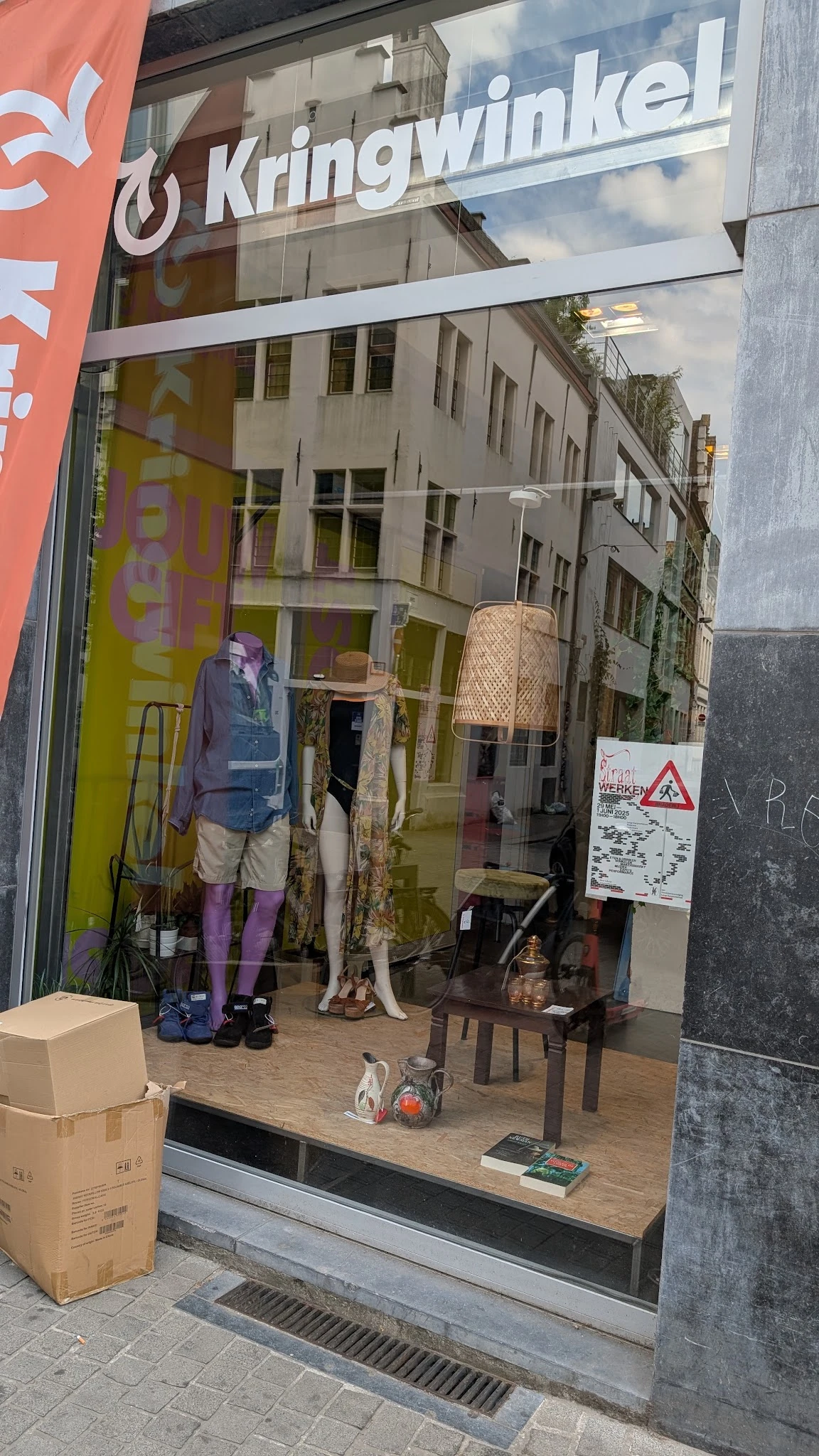 Etalage met tweedehands kleding op mannequins en hangers, inclusief een rieten lamp en accessoires