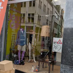 Etalage met tweedehands kleding op mannequins en hangers, inclusief een rieten lamp en accessoires