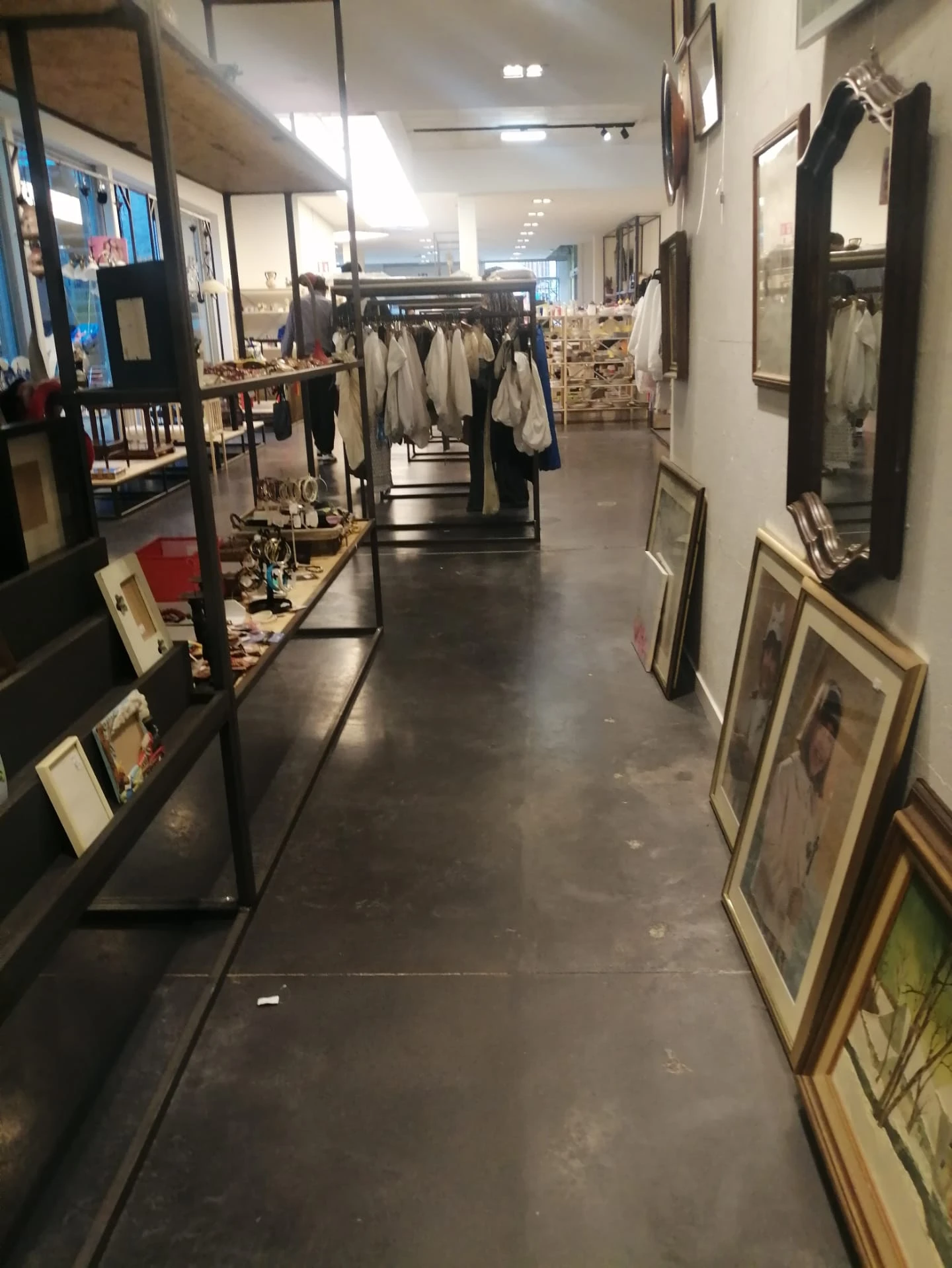 Winkelgang met kledingrekken, afbeeldingen in frames aan de rechterkant en diverse tweedehands spullen