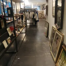 Winkelgang met kledingrekken, afbeeldingen in frames aan de rechterkant en diverse tweedehands spullen