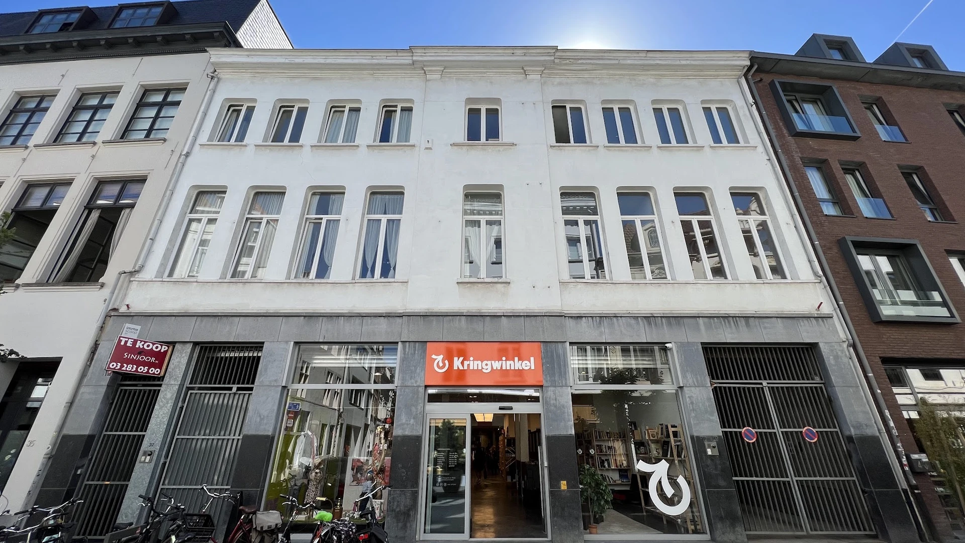 Witte gevel van een kringloopwinkel met rode Kringwinkel-logo op de begane grond en grote winkelramen