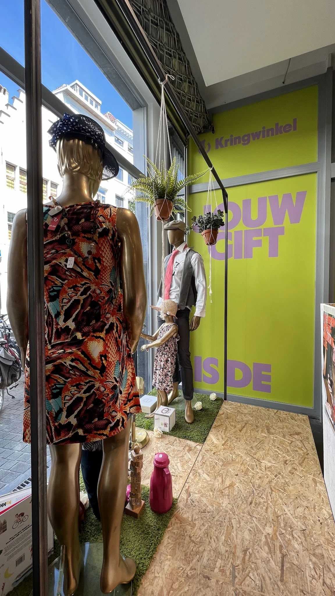 Etalage met tweedehands kleding op manikins, inclusief een felgekleurde jurk en jasje tegen gele wand