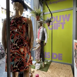 Etalage met tweedehands kleding op manikins, inclusief een felgekleurde jurk en jasje tegen gele wand