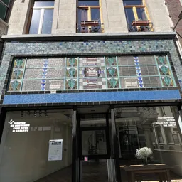 Gevel van kringloopwinkel met decoratieve tegelwerk in blauwe en groene tinten boven de ingang