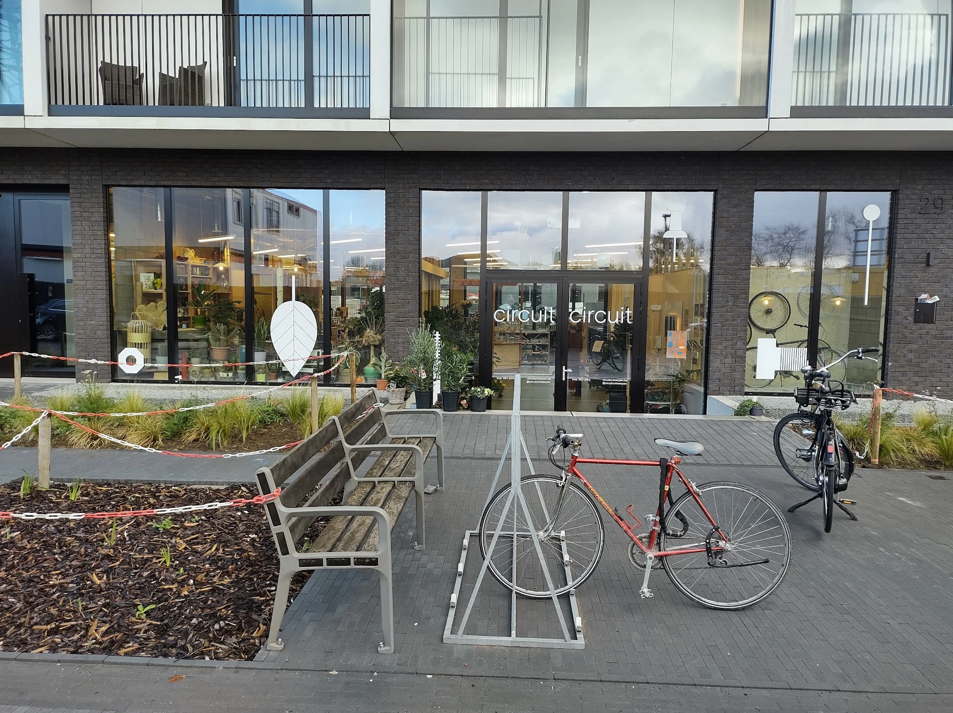 Voorkant van de kringloopwinkel met grote glazen deuren, plantenbakken en fietsenrek op het terras