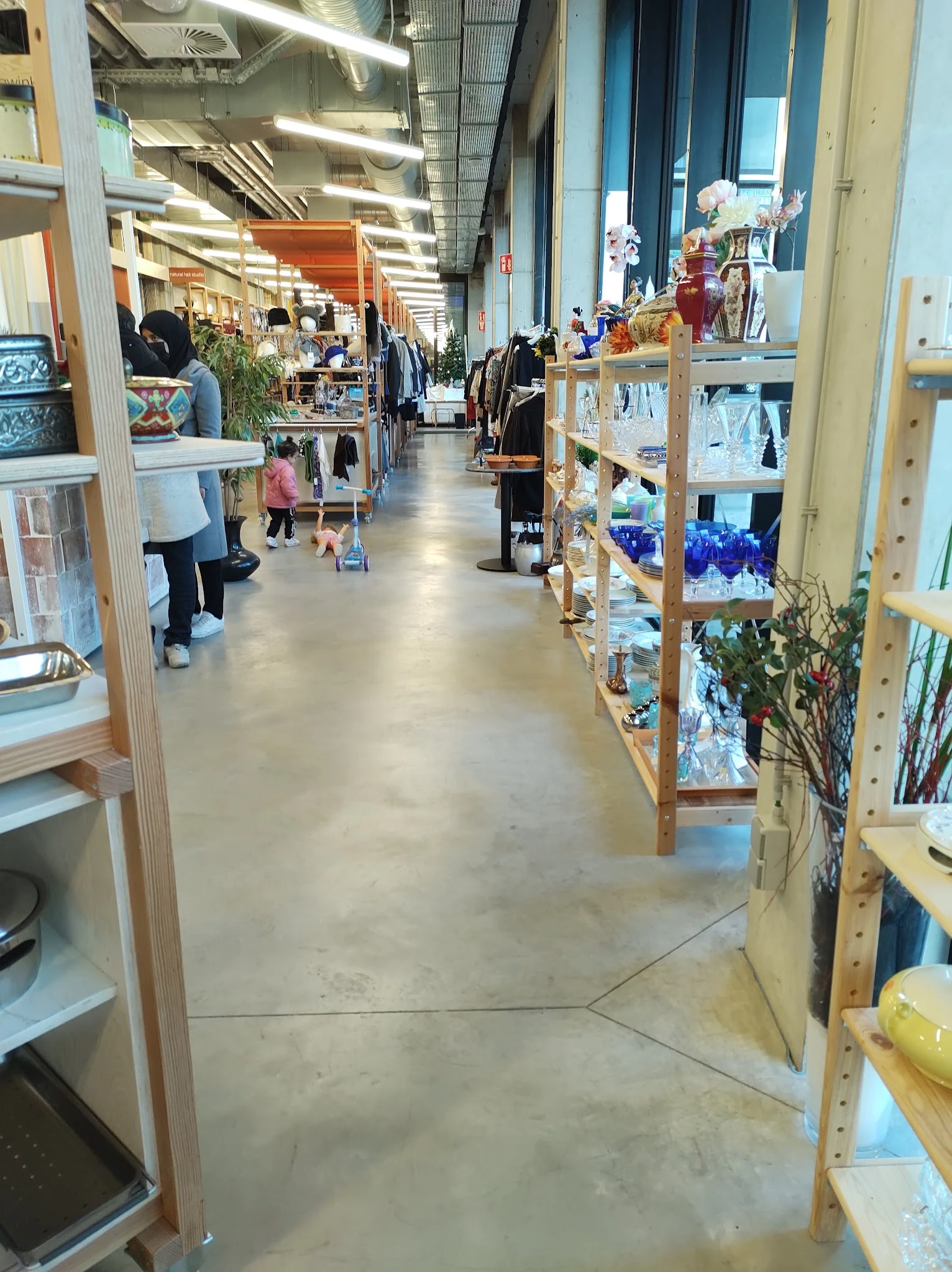 Winkelgang met houten stellingen vol spullen aan beide zijden, enkele bezoekers in de kringloopwinkel