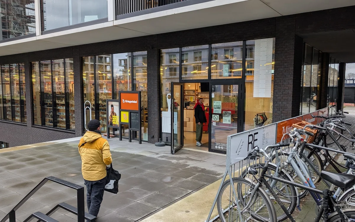 Gevelvoorzijde van kringloopwinkel met grote ramen, oranje logo en ingang, fietsenstalling op straat