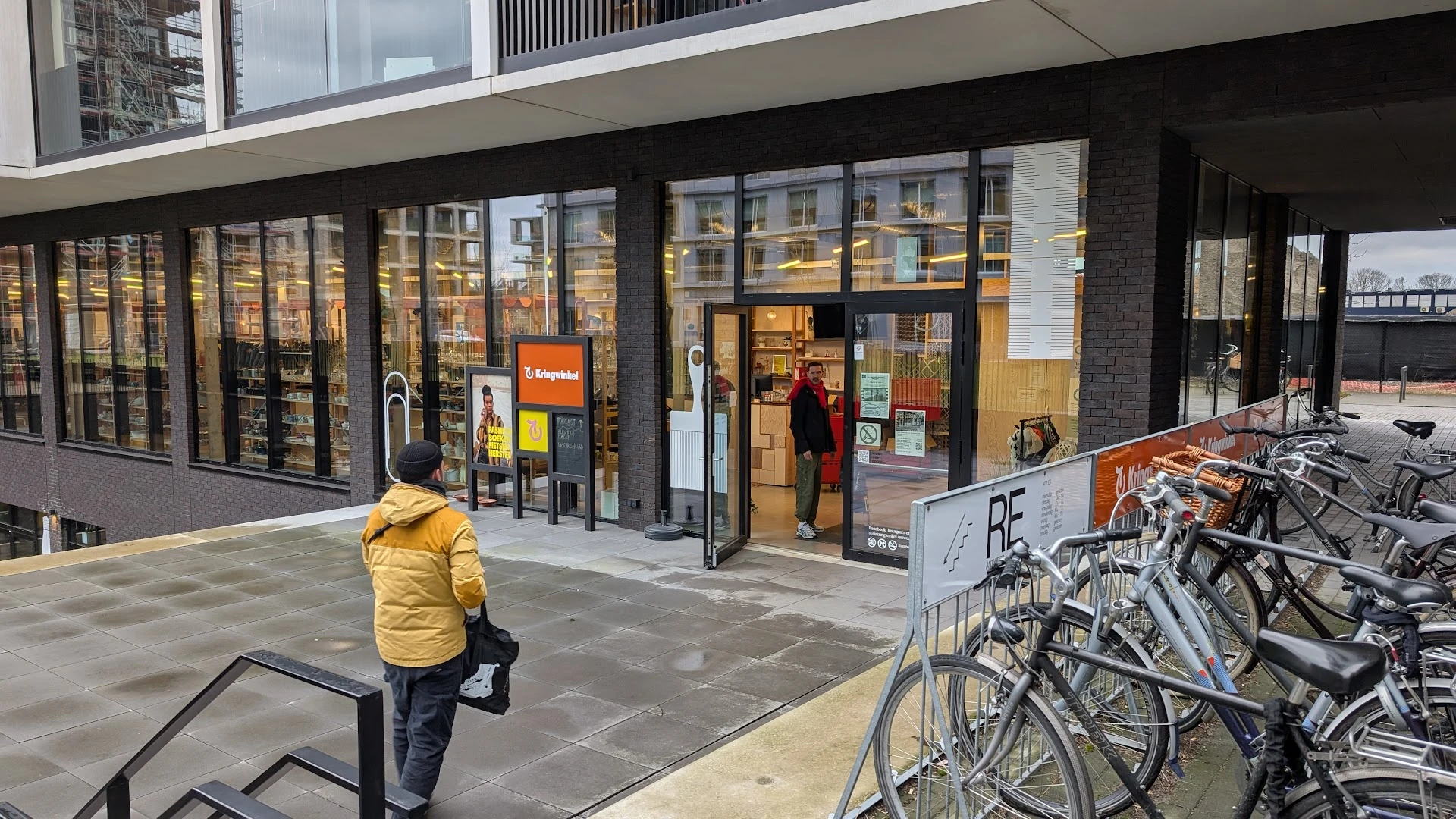 Gevelvoorzijde van kringloopwinkel met grote ramen, oranje logo en ingang, fietsenstalling op straat