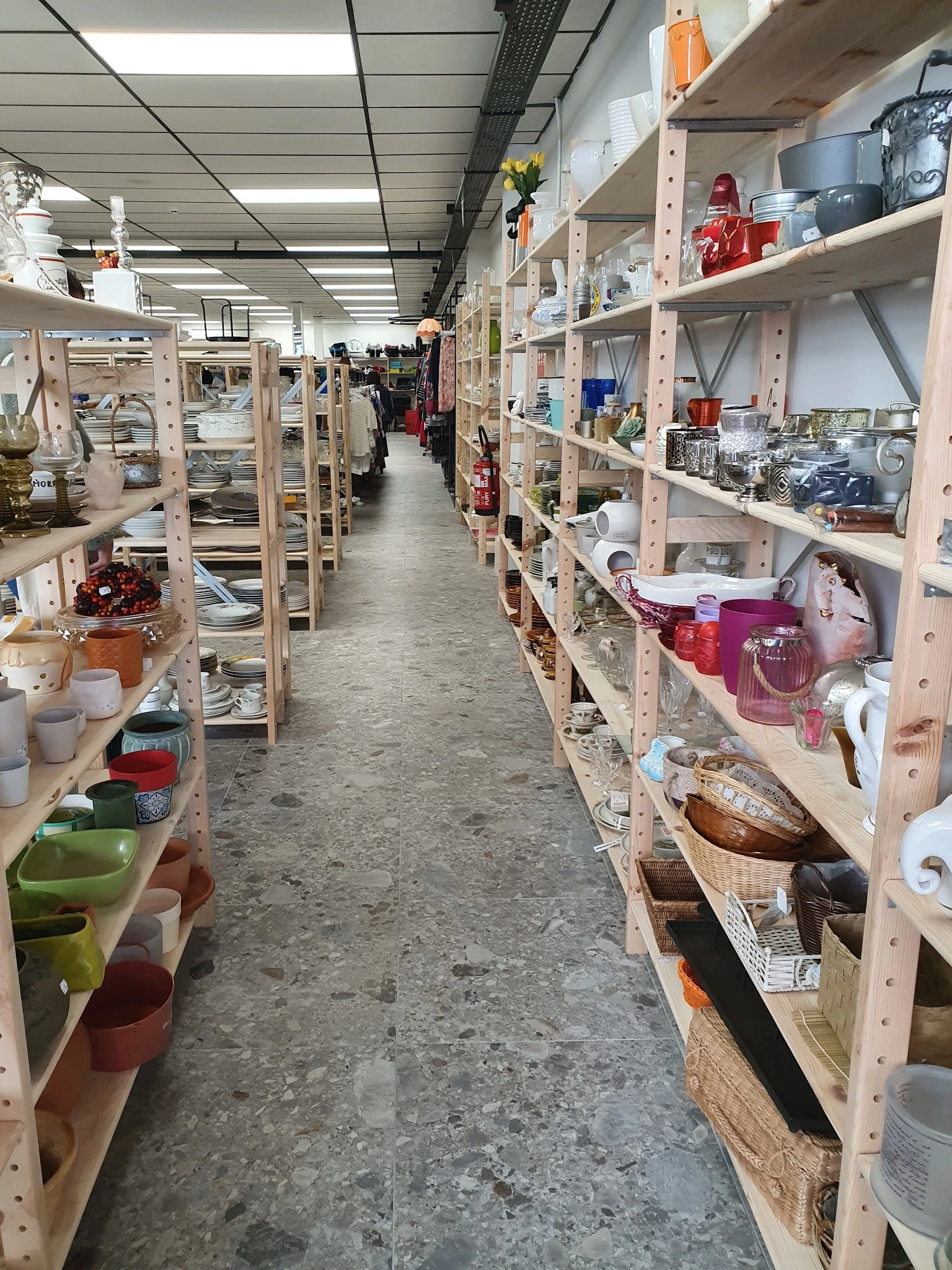 Lange doorgang tussen houten stellingen vol huishoudspullen, vazen, servies en diverse gebruiksartikelen in kringloopwinkel