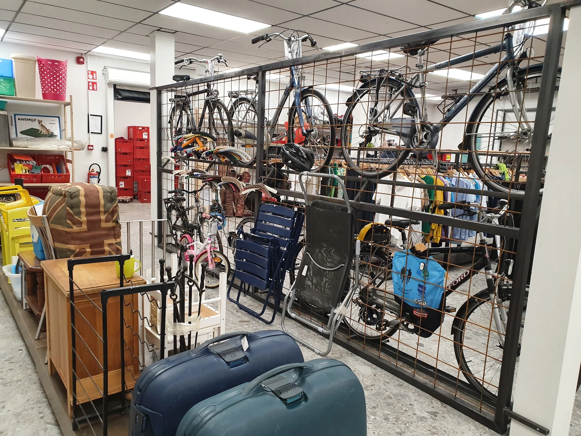 Fietsen en fietsaccessoires uitgestald op een rek achter een gaas in een kringloopwinkel, met daarnaast koffers en huishoudar