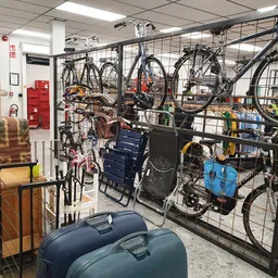 Fietsen en fietsaccessoires uitgestald op een rek achter een gaas in een kringloopwinkel, met daarnaast koffers en huishoudar