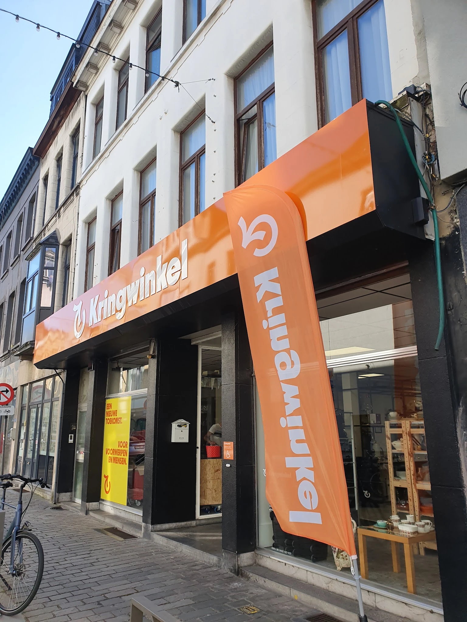 Oranje gevel van kringloopwinkel met grote naam en logo, winkelpui met etalage en ingang zichtbaar