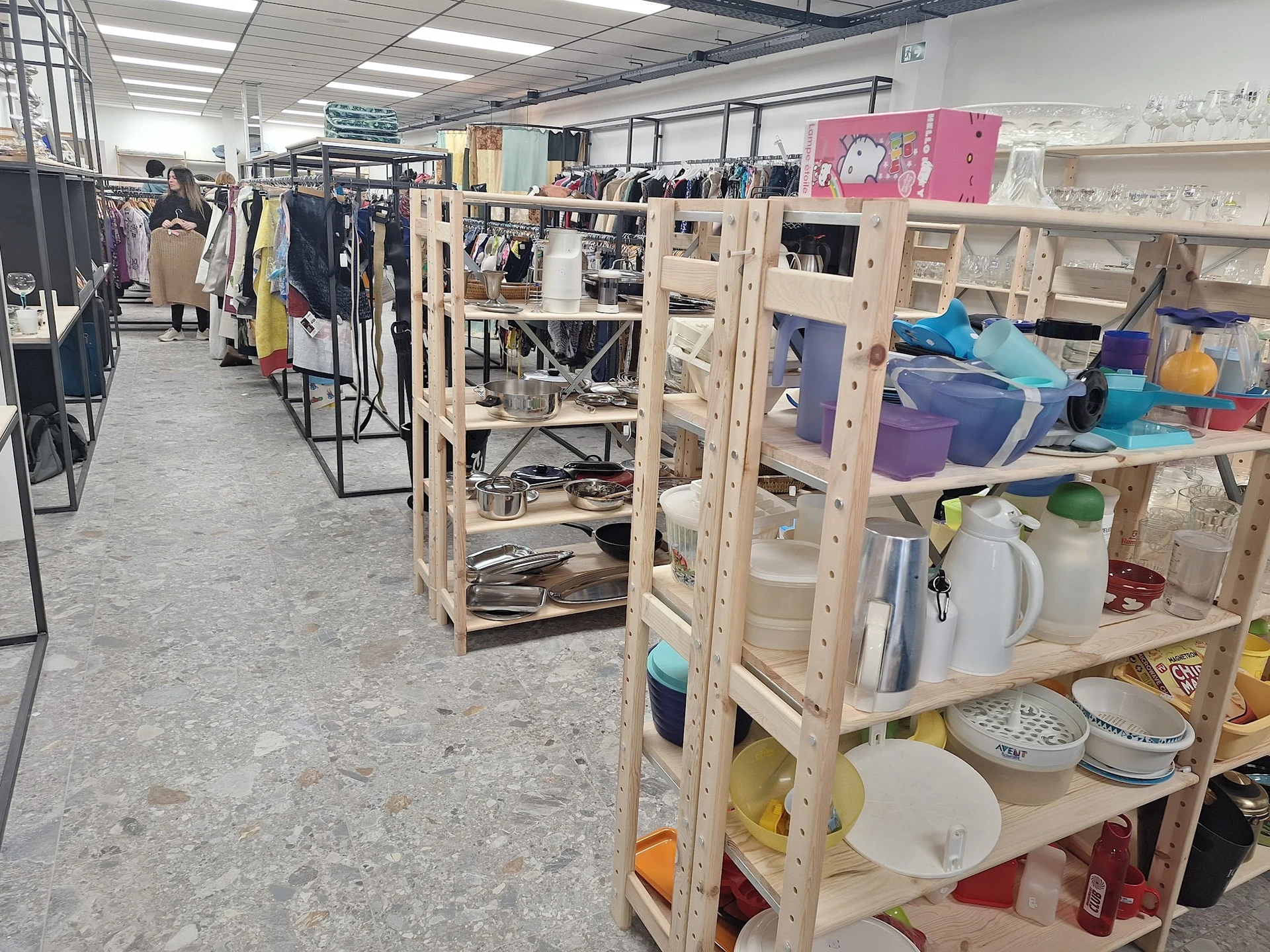 Winkelvloer met houten stellingen vol huishoudelijke spullen, servies en dozen; op de achtergrond kledingrekken en verlichte 