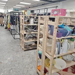 Winkelvloer met houten stellingen vol huishoudelijke spullen, servies en dozen; op de achtergrond kledingrekken en verlichte 