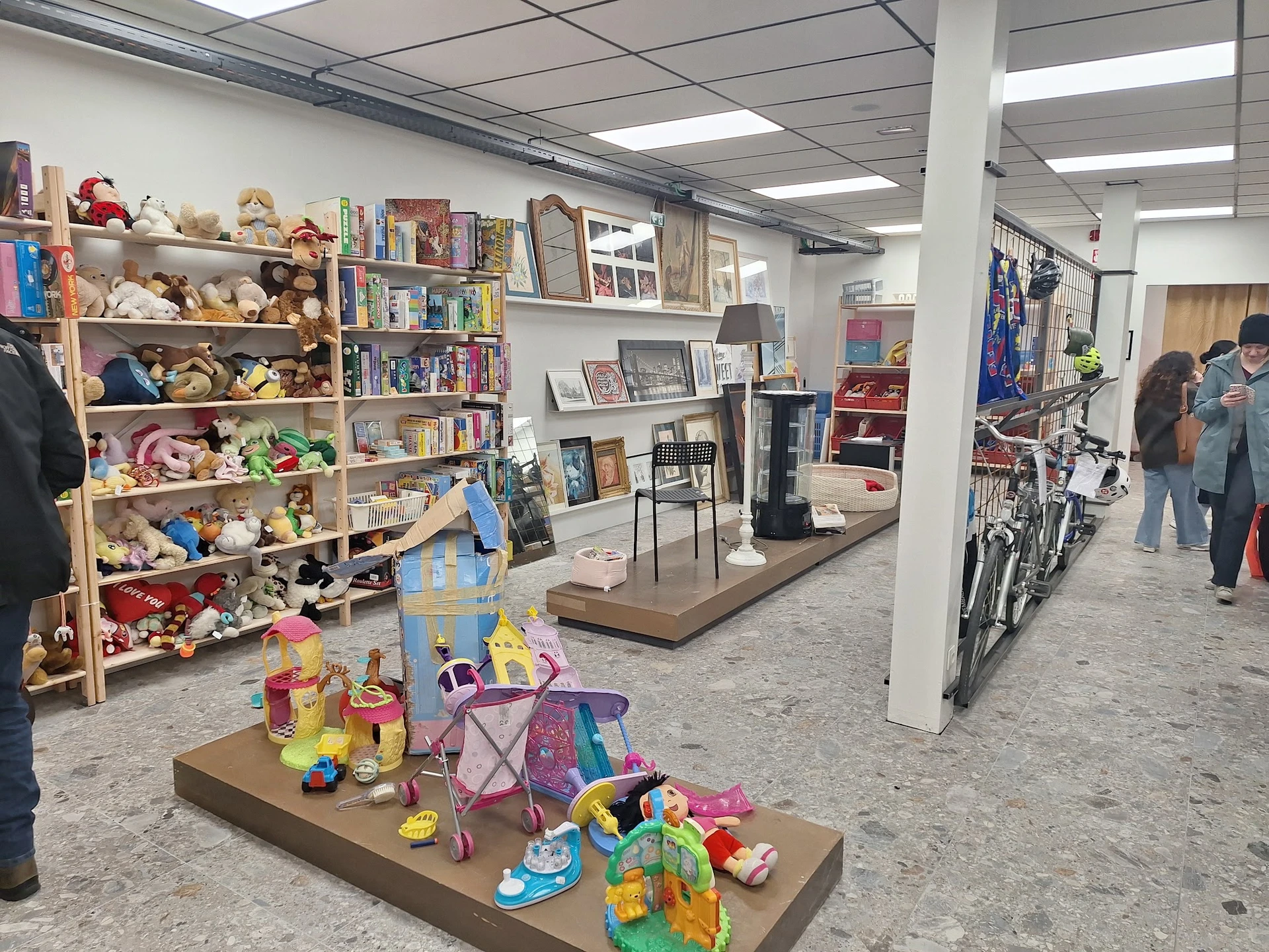 Winkelvloer met speelgoed en boeken op uitstallingstafels, boekenrekken tegen de muur en fietsen op de achtergrond