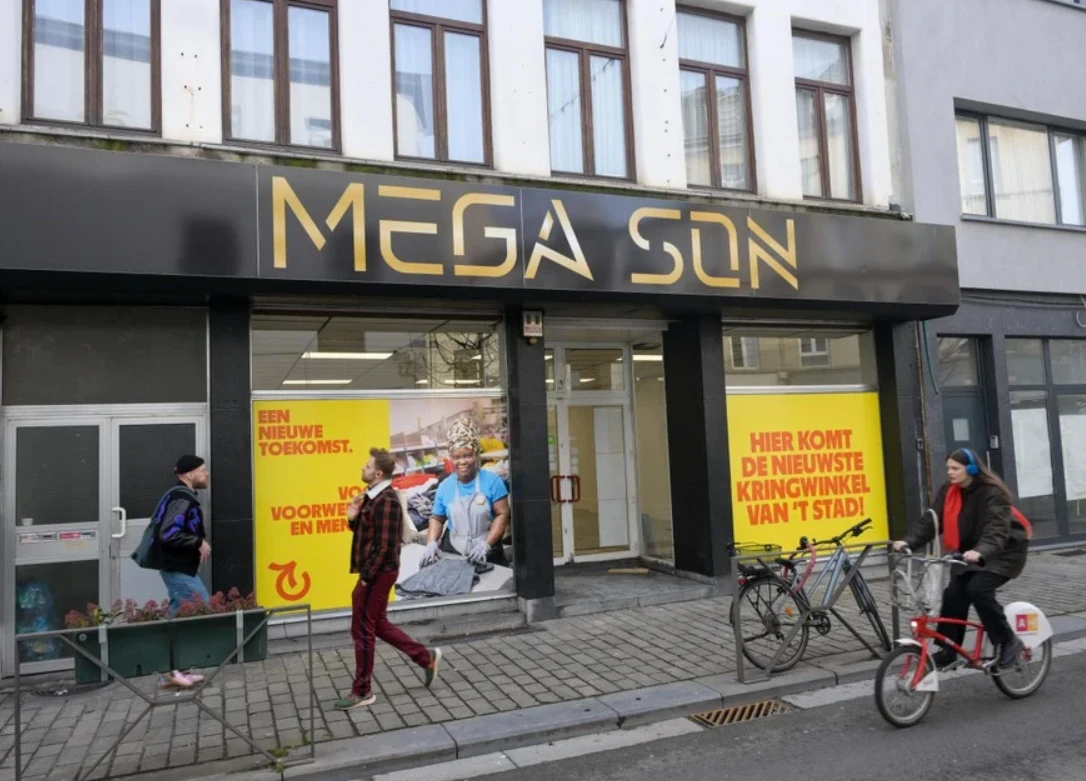 Zwarte gevel van kringloopwinkel Mega Son met gele letters, twee etalageruiten met gele promotieteksten, voetgangers en fiets