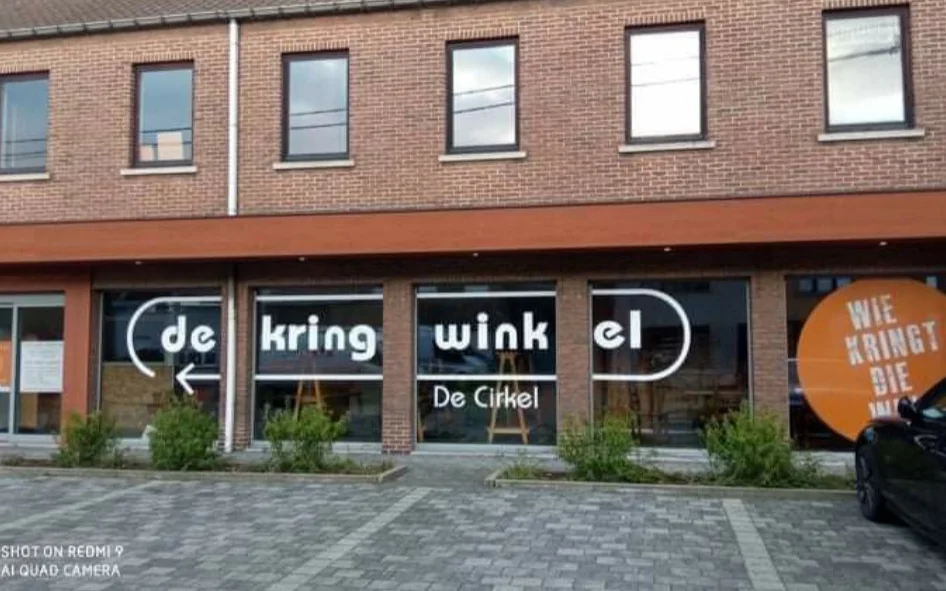 Gevel van kringloopwinkel met bakstenen voorgevel, grote ramen en zwarte uithangtekst met winkelnaam