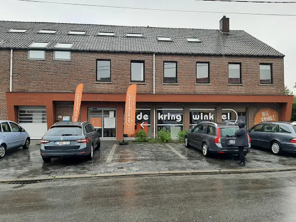 Voorgevel van kringloopwinkel met oranje entree, bakstenen bovenverdieping, parkeerplaats met auto's