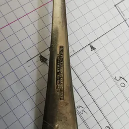 Close-up van een oude metalen buissleutel of spuitbus met ingegraveerde tekst, op ruitjespapier.
